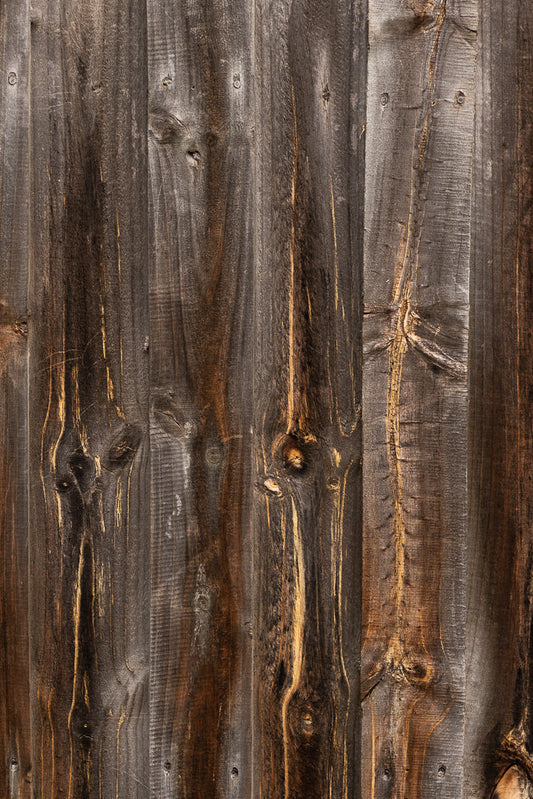 A close-up, vertical shot of weathered, dark brown wooden planks. The planks have visible grain patterns, knots, and some lighter brown or golden streaks. Several nails are visible, securing the planks.