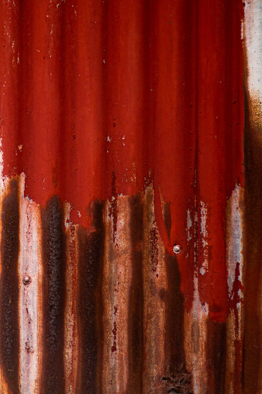 A close-up, abstract view of weathered, corrugated metal with vertical ridges. The surface is predominantly a deep, rusty red, with areas of peeling paint revealing lighter metal underneath. Streaks of rust and water stains run down the surface, creating a textured, aged appearance. Some small bubbles or imperfections are visible on the surface.