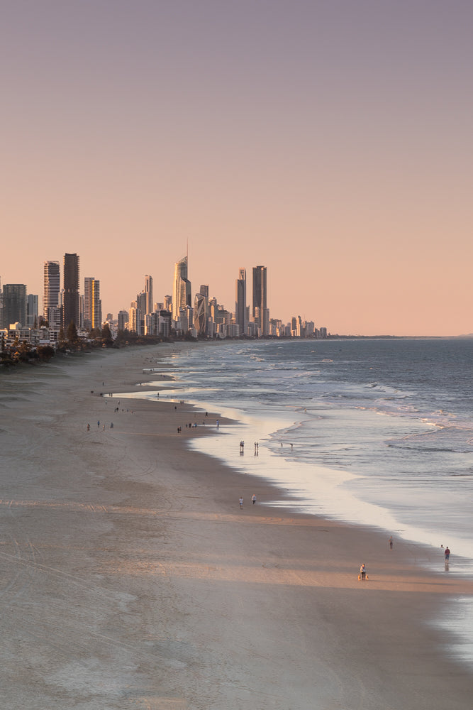 A wide shot of a beach with a city skyline in the background during sunset. The sky is a soft pink and purple, and the ocean has gentle waves. Several people are walking on the beach and in the water.
