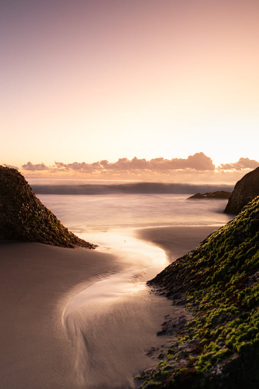 A serene beach scene at sunset, with soft pastel colors in the sky. The gentle waves create a blurred, silky effect on the water. Two moss-covered rocks frame a sandy inlet, reflecting the warm, golden light of the setting sun.