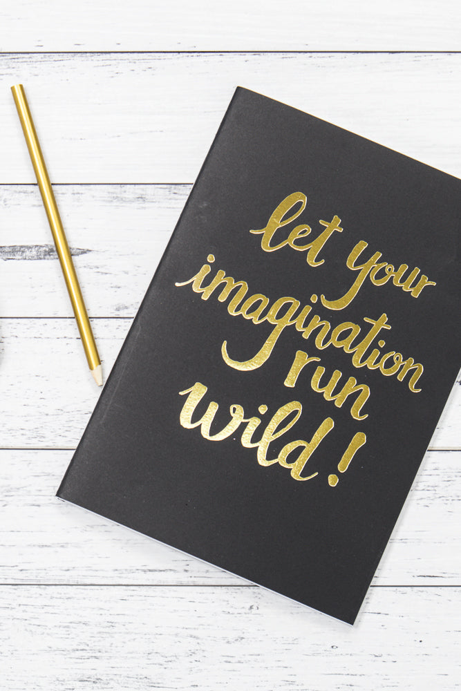 A black notebook with gold lettering that reads "let your imagination run wild!" lies on a white wooden surface next to a gold pencil.
