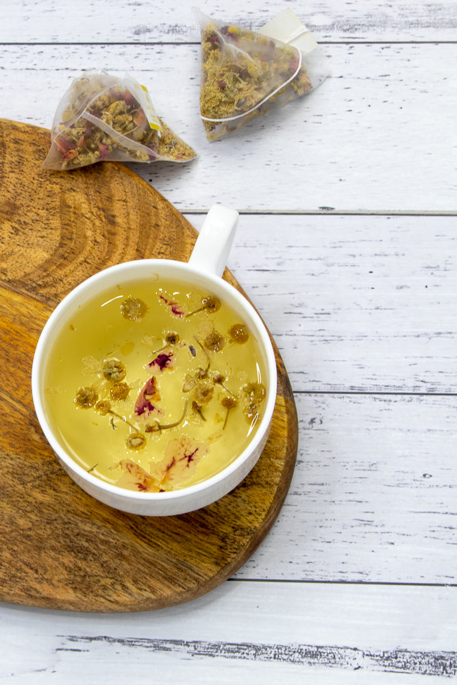 A white mug filled with golden herbal tea, containing visible chamomile flowers and rose petals, sits on a wooden board. Two pyramid-shaped tea bags filled with dried herbs and flowers are placed on a white wooden surface behind the mug.