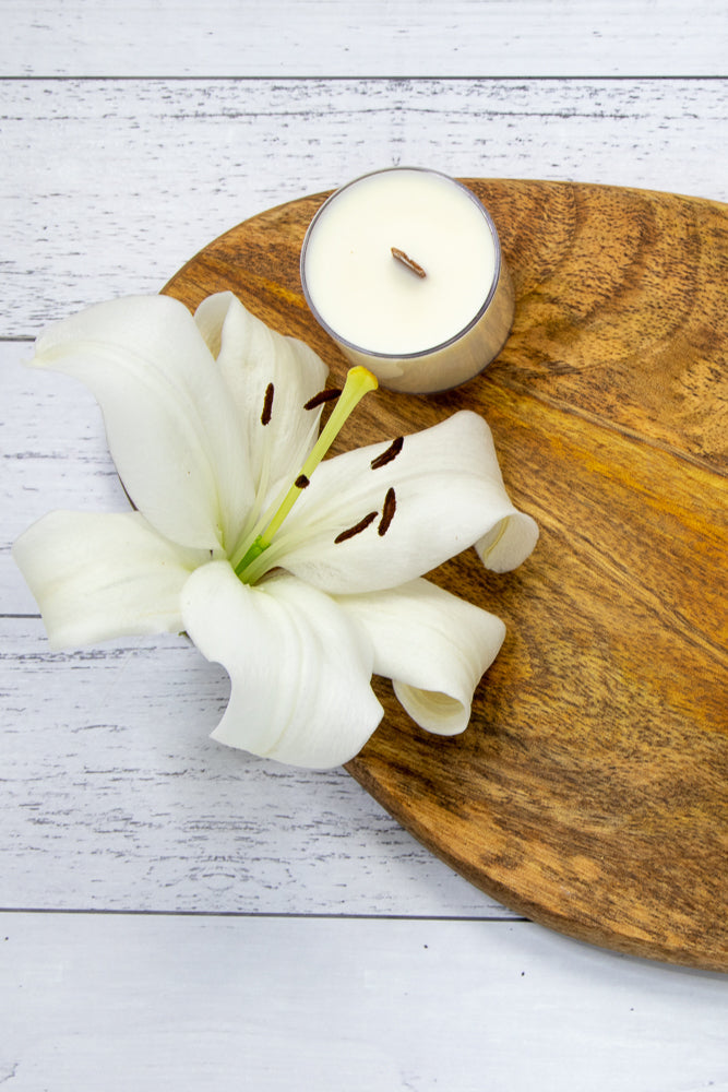 A white mug filled with golden herbal tea, containing visible chamomile flowers and rose petals, sits on a wooden board. Two pyramid-shaped tea bags filled with dried herbs and flowers are placed on a white wooden surface behind the mug.