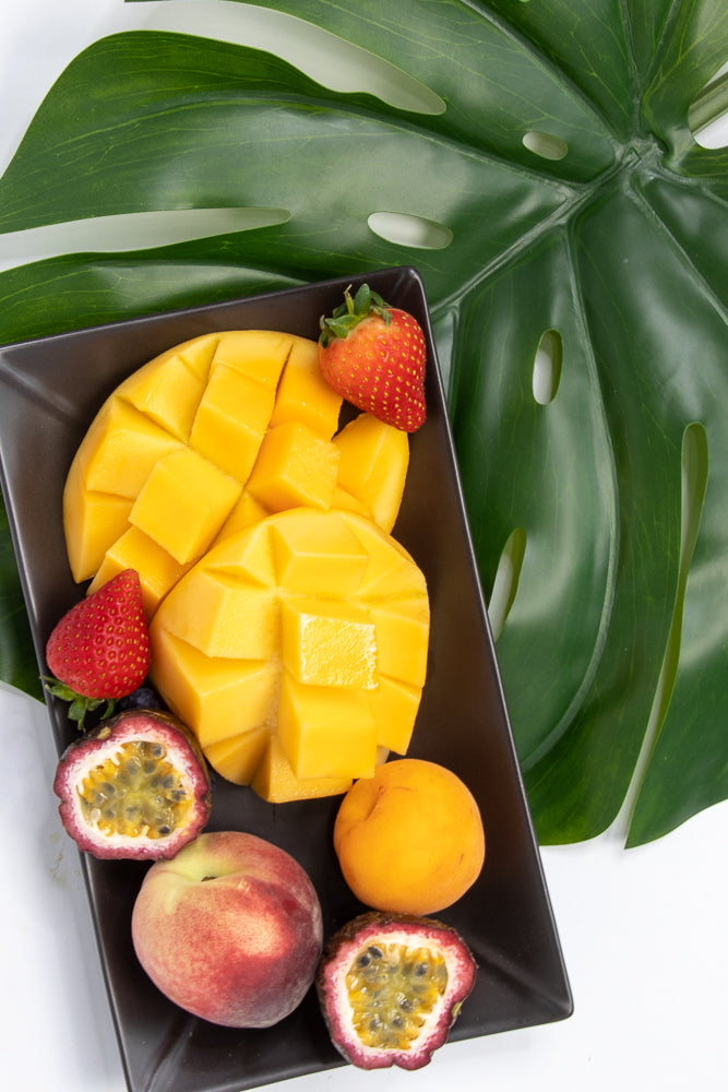 A dark square plate holds a variety of fresh fruits, including cubed mango, whole strawberries, a halved passion fruit, a peach, and an apricot, all arranged on a white surface with a large green monstera leaf in the background.