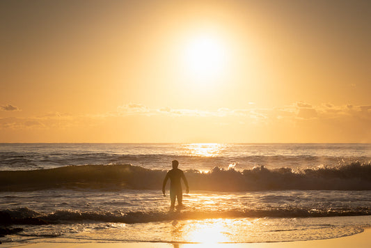 A surfer stands in the ocean waves at sunrise, silhouetted against the bright orange sun.