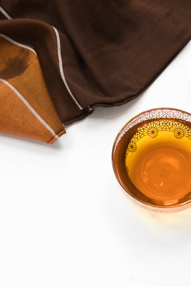 A close-up overhead shot shows a glass of amber liquid with a decorative pattern on the rim, next to a folded brown and orange striped cloth on a white background.