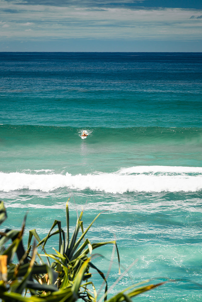 A surfer paddles through a turquoise wave under a cloudy sky. The foreground is blurred with green foliage.