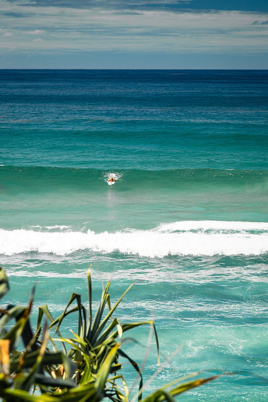 A surfer paddles through a turquoise wave under a cloudy sky. The foreground is blurred with green foliage.