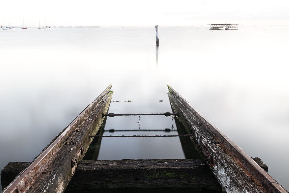 A long exposure shot of an old wooden boat slipway leading into calm, misty water. Sailboats and a distant structure are visible on the horizon under a bright, overcast sky.