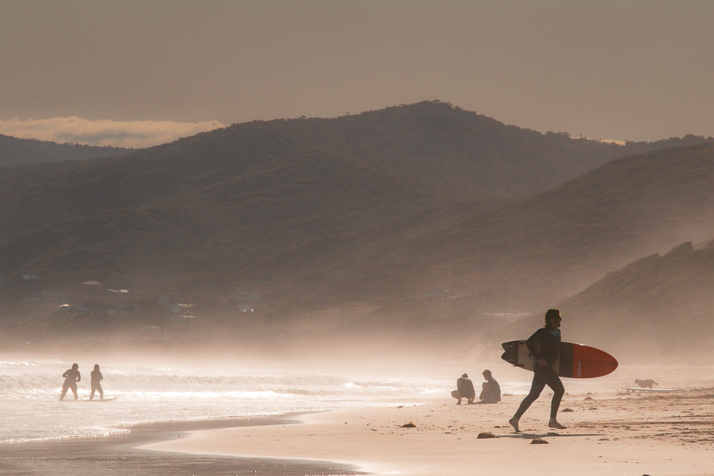 A surfer in a wetsuit runs along a sandy beach carrying a surfboard. In the background, two people stand in the ocean waves, and two others sit on the sand. Mountains are visible in the distance under a hazy sky.