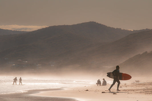 A surfer in a wetsuit runs along a sandy beach carrying a surfboard. In the background, two people stand in the ocean waves, and two others sit on the sand. Mountains are visible in the distance under a hazy sky.