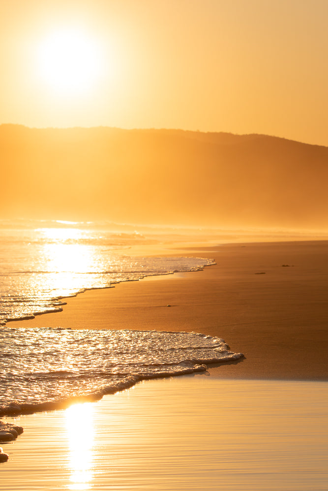 Golden sunset over a calm beach with gentle waves lapping the shore. The sun reflects brightly on the wet sand and the water, creating a warm, serene atmosphere.