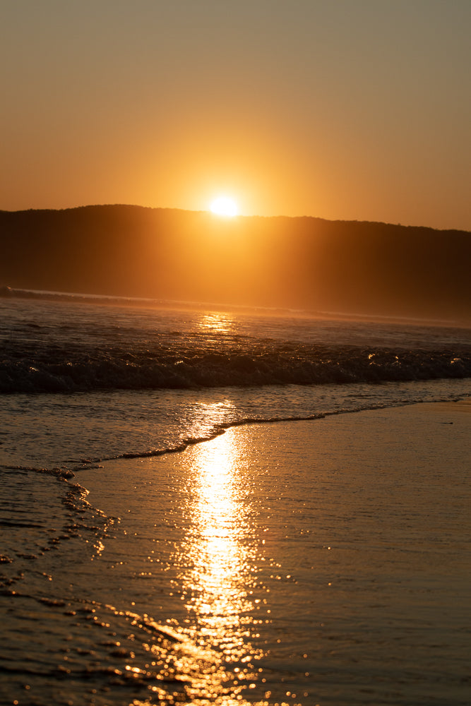 The sun sets behind a silhouetted hill, casting a golden glow over the ocean waves and wet sand. The water reflects the warm light, creating a shimmering path towards the shore.