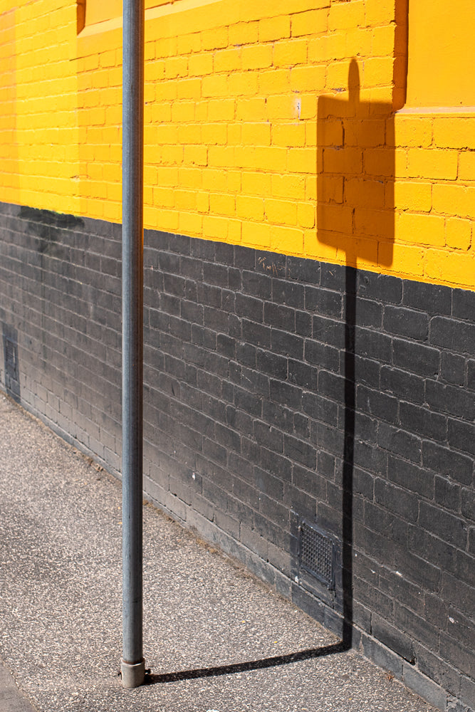 A grey metal pole stands in front of a brick wall painted with a horizontal yellow stripe above a dark grey stripe. A shadow of a sign is cast on the wall.