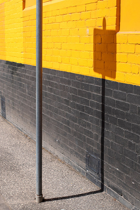 A grey metal pole stands in front of a brick wall painted with a horizontal yellow stripe above a dark grey stripe. A shadow of a sign is cast on the wall.