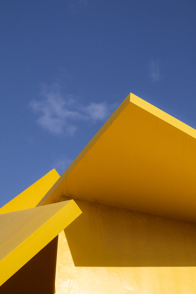 Abstract geometric shapes of bright yellow against a clear blue sky with a few wispy clouds. The image focuses on the sharp angles and planes of the structure, creating a minimalist and modern composition.