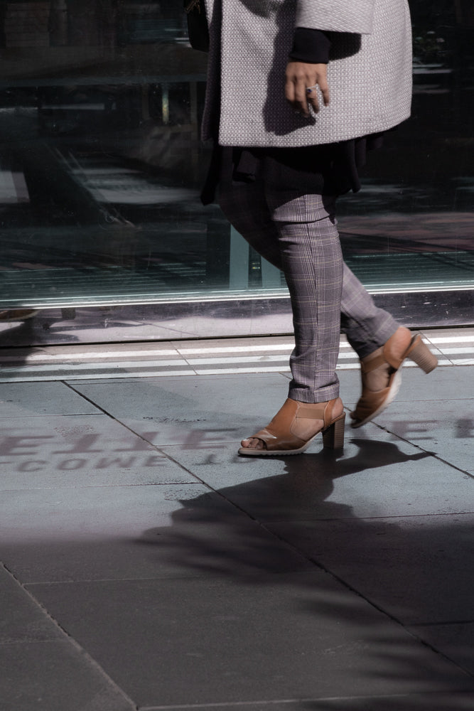 A person walks on a sidewalk in front of a reflective glass building. They are wearing a grey patterned coat, grey plaid pants, and tan open-toed heels. The sun casts a shadow of the person on the ground.