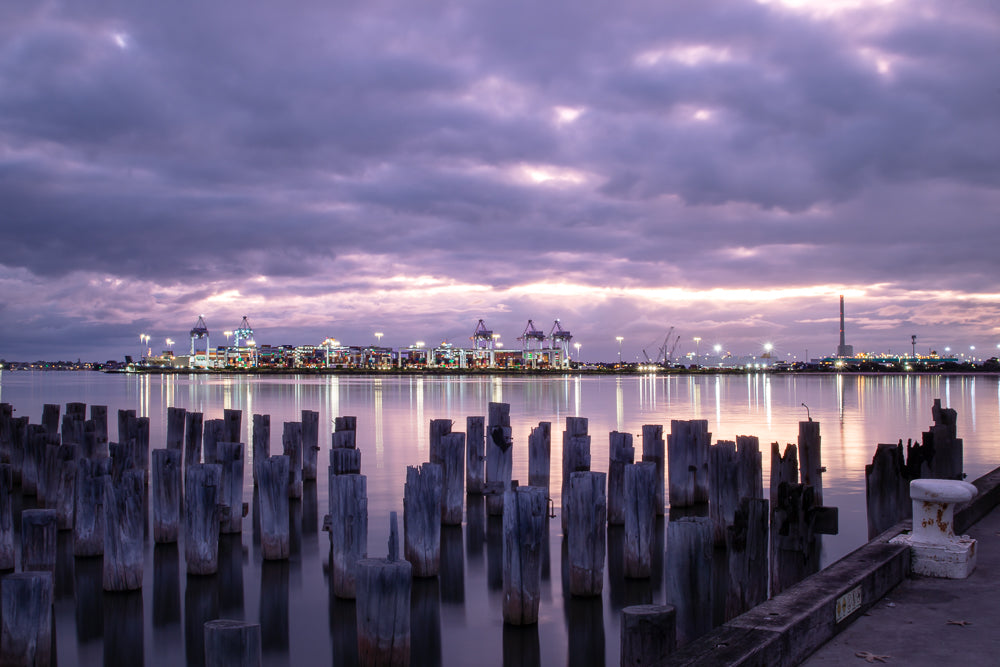 A twilight view of a harbor with weathered wooden pilings in the foreground and a brightly lit industrial port with cranes in the background under a cloudy purple sky.