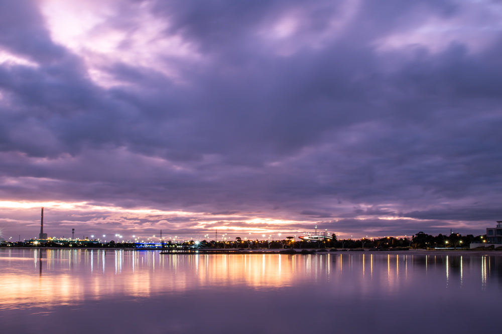 A serene twilight scene with a purple and pink sky reflected in calm water. Lights from a distant city skyline twinkle along the horizon, creating shimmering reflections.