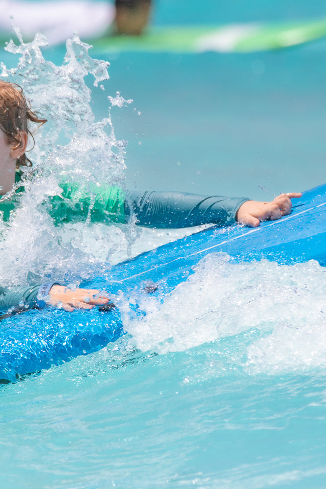 A child with brown hair wearing a green rash guard is being splashed with water while holding onto a blue surfboard.