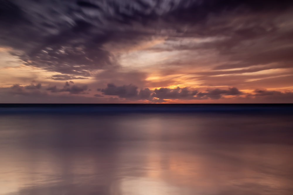 A long exposure shot of a sunset over the ocean. The sky is filled with dramatic clouds in shades of purple, orange, and pink, with the sun peeking through the clouds. The ocean is calm and reflects the colors of the sky.
