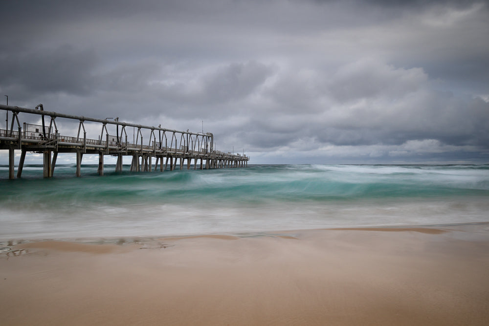 A long pier extends over the ocean under a cloudy sky. The water below the pier is a vibrant turquoise, with waves blurred by a long exposure, creating a sense of movement. The foreground shows a sandy beach.