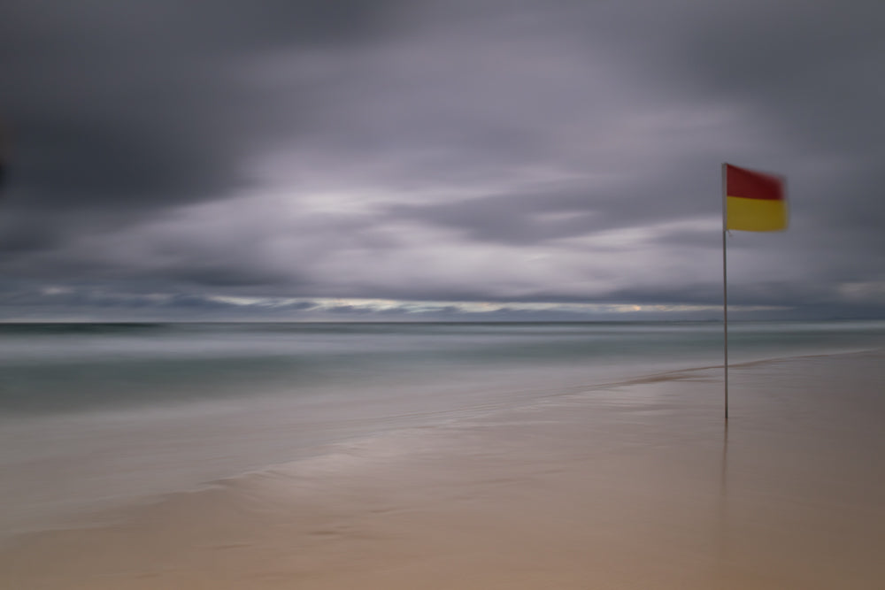 A red and yellow flag on a pole stands on a sandy beach under a cloudy sky. The ocean is calm and the waves are blurred, creating a serene, atmospheric effect.