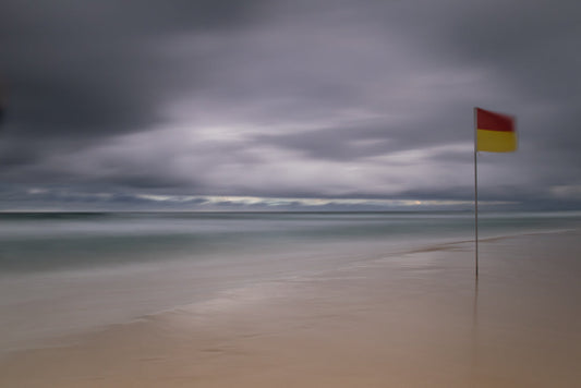 A red and yellow flag on a pole stands on a sandy beach under a cloudy sky. The ocean is calm and the waves are blurred, creating a serene, atmospheric effect.