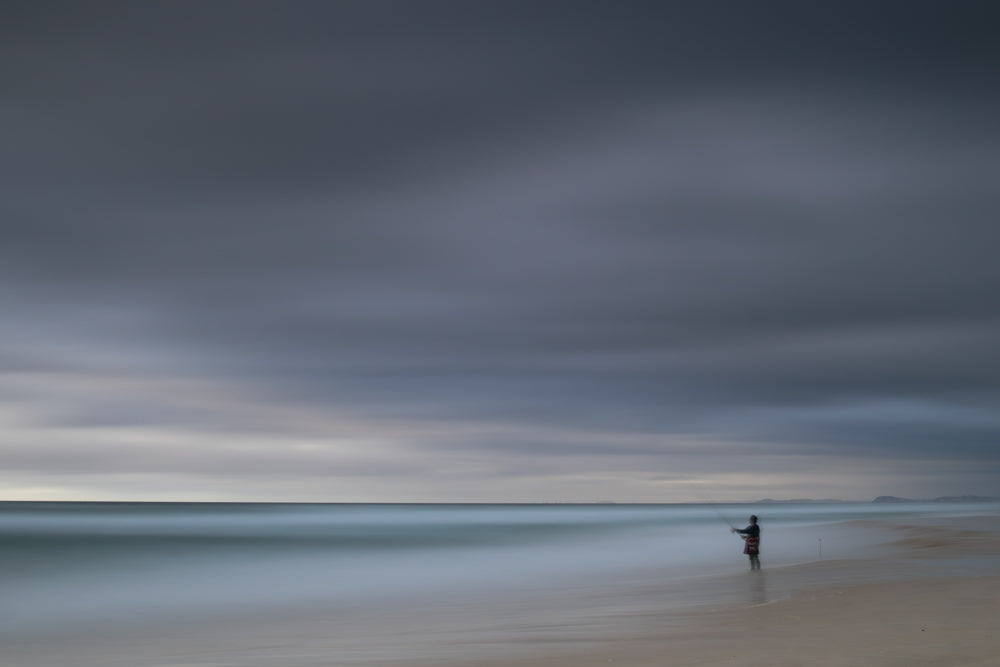 A lone figure stands on a sandy beach, fishing in the ocean under a dramatic, cloudy sky. The long exposure blurs the water and clouds, creating a serene and atmospheric scene.