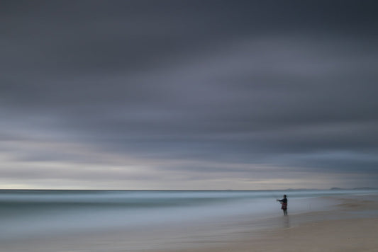 A lone figure stands on a sandy beach, fishing in the ocean under a dramatic, cloudy sky. The long exposure blurs the water and clouds, creating a serene and atmospheric scene.