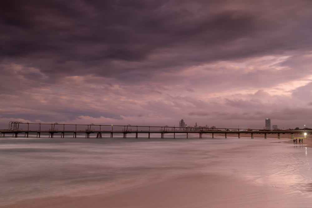 A long pier extends over the ocean under a dramatic, cloudy sky at dusk. The water is calm and reflects the soft light, creating a serene atmosphere. A few people are visible on the beach near the pier's end.
