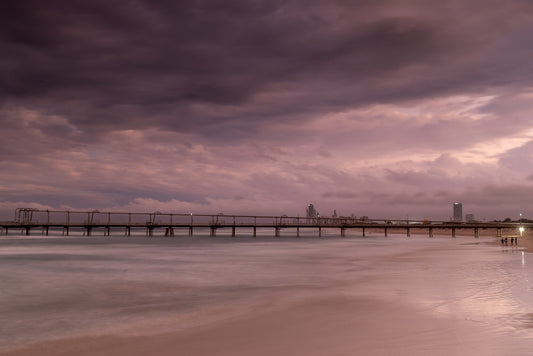 A long pier extends over the ocean under a dramatic, cloudy sky at dusk. The water is calm and reflects the soft light, creating a serene atmosphere. A few people are visible on the beach near the pier's end.