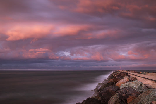A lighthouse stands at the end of a rocky pier as dramatic pink and purple clouds fill the sky during sunset over the ocean.