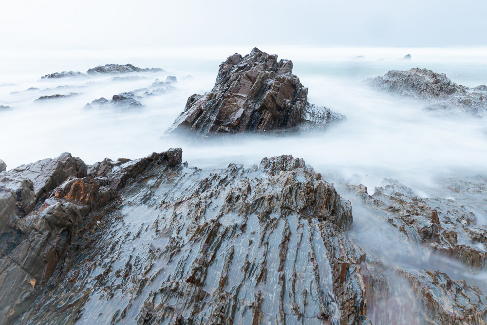 A long exposure shot of jagged, wet rocks on a coastline, with misty water swirling around them. The rocks are dark brown and grey, with some reddish-brown hues. The water is a soft, ethereal white, creating a dreamlike atmosphere.