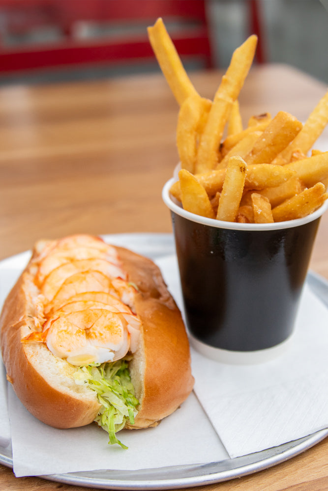 A lobster roll filled with lobster meat and lettuce sits on a metal tray next to a cup of french fries.