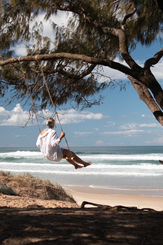 A person wearing a white hoodie and blue bikini bottoms sits on a rope swing, looking out at the ocean. The swing is suspended from a tree branch, and the beach is visible in the foreground.
