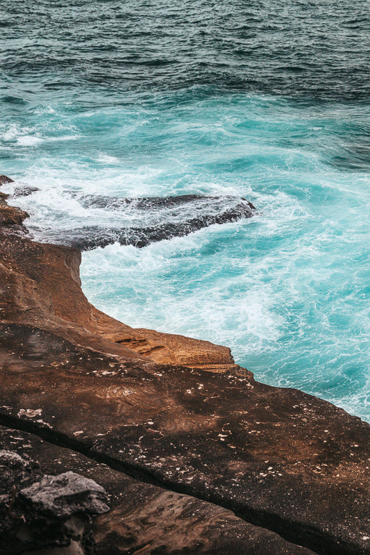 A close-up view of turquoise ocean waves crashing against dark, textured rocks. The water is a vibrant blue-green, with white foam swirling around the rocky shore.