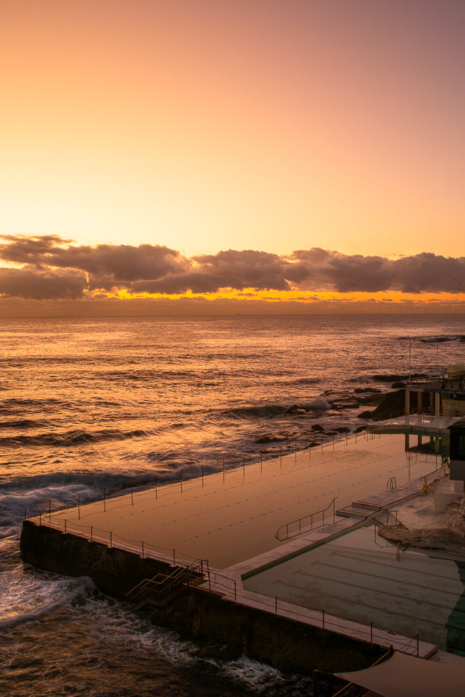 A wide shot of an ocean pool at sunrise. The sky is a gradient of orange and yellow, with scattered clouds. The ocean is a deep orange, with waves crashing against the rocks surrounding the pool. The pool itself is filled with calm, reflective water.