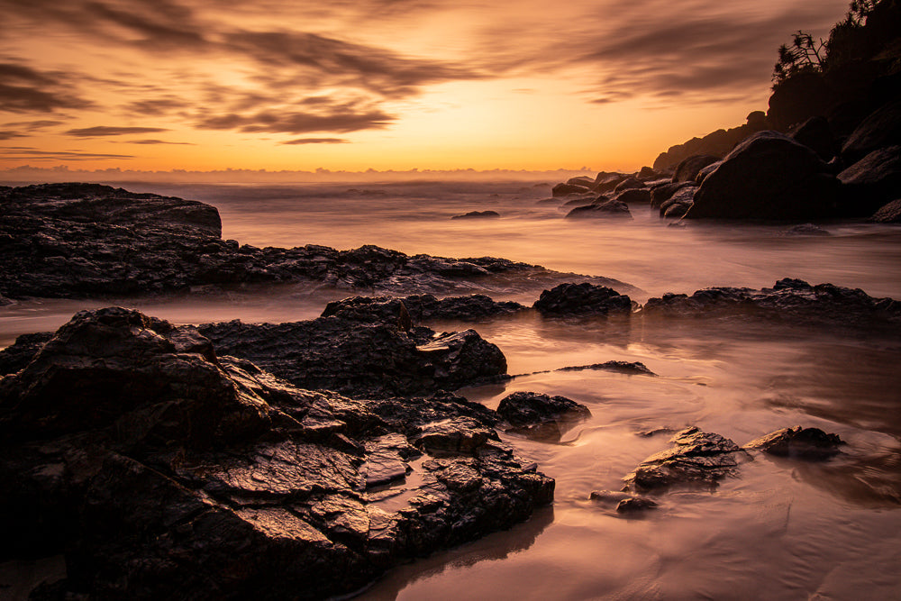 A dramatic sunset over a rocky coastline. The sky is filled with fiery orange and golden clouds, casting a warm glow on the ocean and wet rocks. The water appears smooth and misty due to a long exposure, creating a serene and ethereal atmosphere.