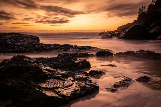 A dramatic sunset over a rocky coastline. The sky is filled with fiery orange and golden clouds, casting a warm glow on the ocean and wet rocks. The water appears smooth and misty due to a long exposure, creating a serene and ethereal atmosphere.