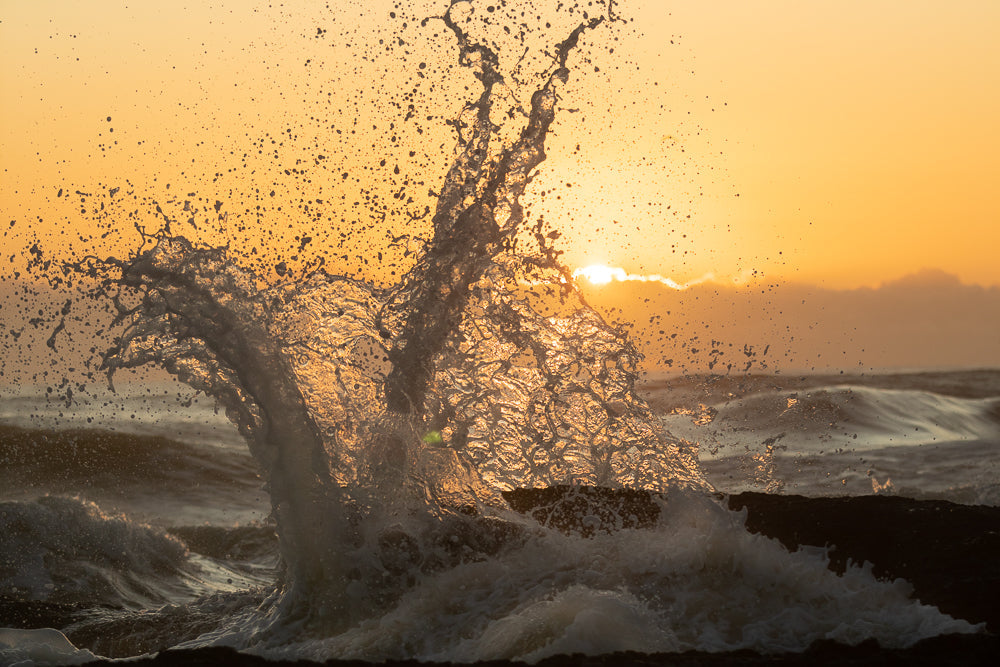 A dramatic wave crashes against dark rocks at sunset. The golden light of the setting sun illuminates the spray, creating a sparkling effect against the orange sky.