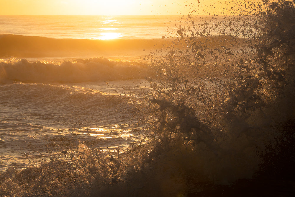 A wave crashes in the foreground, with the sun setting over the ocean in the background. The water is illuminated by the golden light of the sunset, creating a dramatic and beautiful scene.