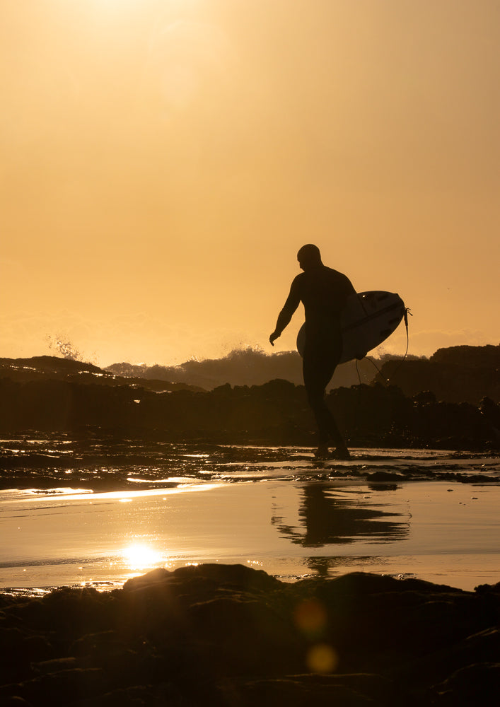 Silhouette of a surfer in a wetsuit carrying a surfboard, walking on wet rocks towards the ocean at sunset. The sun reflects on the water and wet surfaces, creating a warm, golden glow.