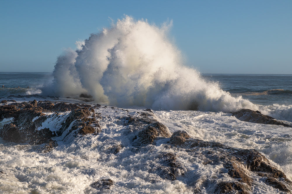 A massive wave crashes against a rocky shore, sending white spray high into the air under a clear blue sky. The turbulent water churns around the dark, wet rocks in the foreground.