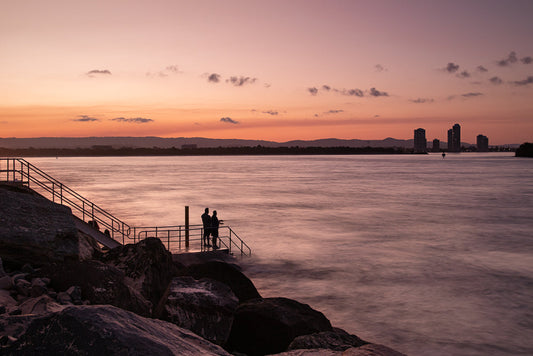 Silhouettes of two people stand on a pier at sunset, looking out at the ocean. A city skyline is visible in the distance. The sky is a gradient of orange and purple, with scattered clouds.