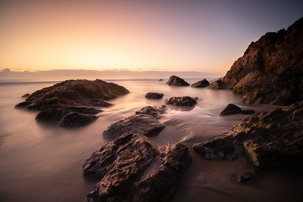 A serene seascape captures the soft glow of a sunset over a rocky coastline. The gentle waves create a silky, blurred effect as they wash over the dark, textured rocks and wet sand, highlighting the tranquil beauty of the ocean at dusk.