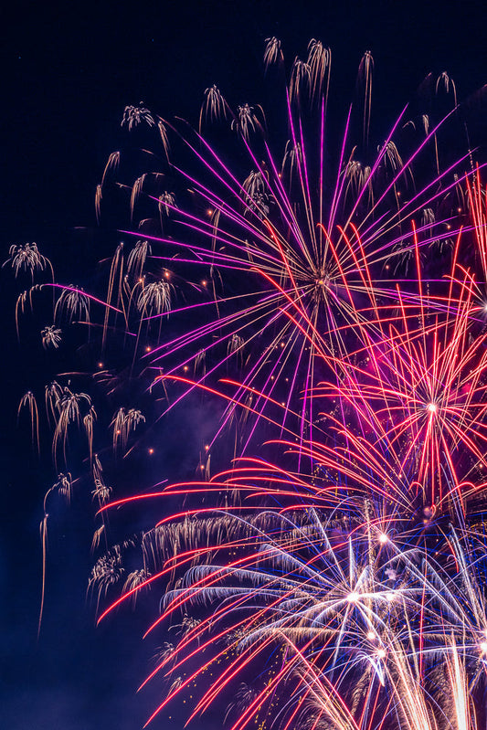 A vibrant display of fireworks illuminates the night sky with streaks of red, pink, and white light against a dark blue background.