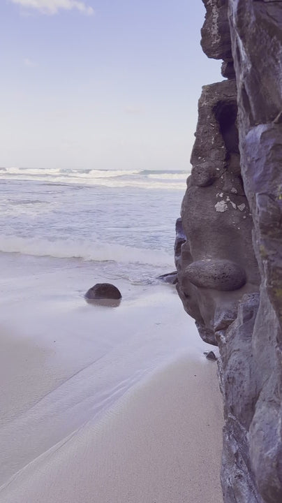 Gentle ocean waves softly washing sandy beach alongside rocky cliff, sunlight creating serene coastal landscape with tranquil water movement