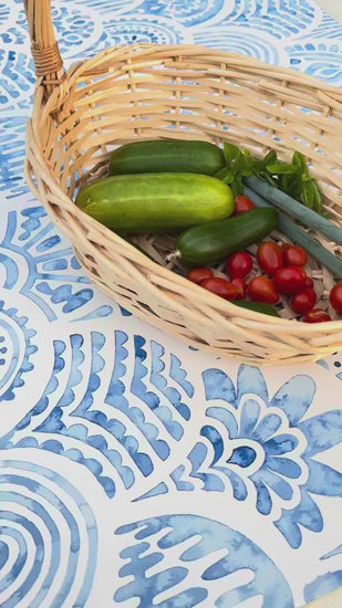 Wicker basket full of fresh vegetables, including cucumbers, ripe cherry tomatoes, and basil leaves, slowly approaching on a patterned tabletop
