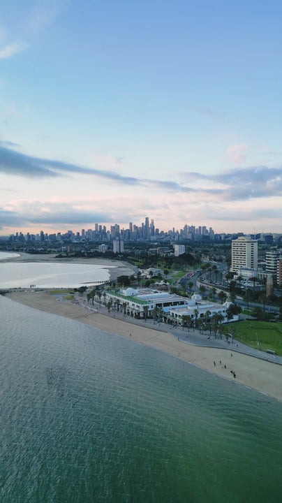 Aerial pull out from Melbourne city skyline and St. Kilda revealing blue calm water of Port Phillip Bay at sunset with pink and blue sky